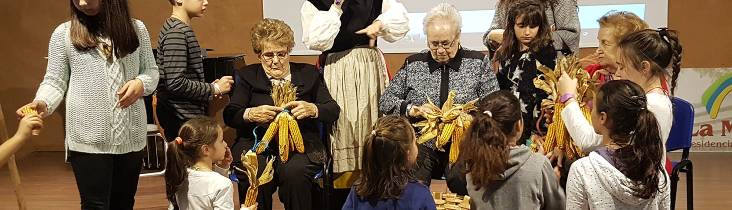 Varios niños y ancianos participan de una actividad de oficios tradicionales dentro del programa de convivencia intergeneracional "Vivencias". Con la temática del campo, tienen en sus manos unas mazorcas de maíz.